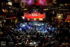 Eine große Menschenmenge, die auf Stühlen vor einer Bühne mit Bildschirmen und Bannern sitzt, die von Scheinwerfern beleuchtet wird und von Geländern umgeben ist und gespannt auf den Beginn eines Online-Marketing-Rockstars-Events wartet, mit einem Wasserzeichen in der linken unteren Ecke.