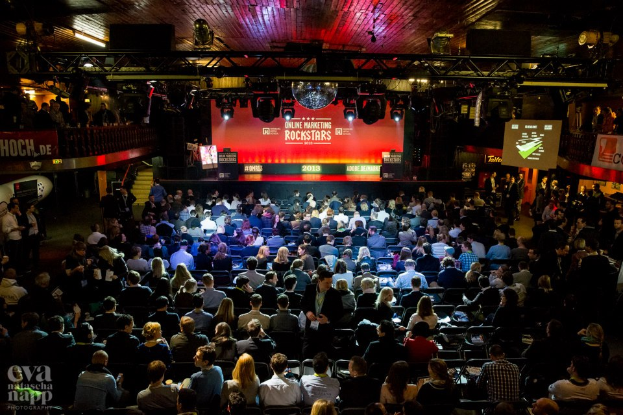 Eine große Menschenmenge, die auf Stühlen vor einer Bühne mit Bildschirmen und Bannern sitzt, die von Scheinwerfern beleuchtet wird und von Geländern umgeben ist und gespannt auf den Beginn eines Online-Marketing-Rockstars-Events wartet, mit einem Wasserzeichen in der linken unteren Ecke.