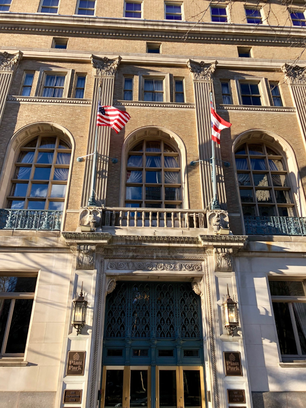 Großes Gebäude mit Glasfront, mehreren Säulen, Geländern und beleuchteter Beschilderung, identifiziert als Plaza Hotel in New York City, vor einem Himmel-Hintergrund.