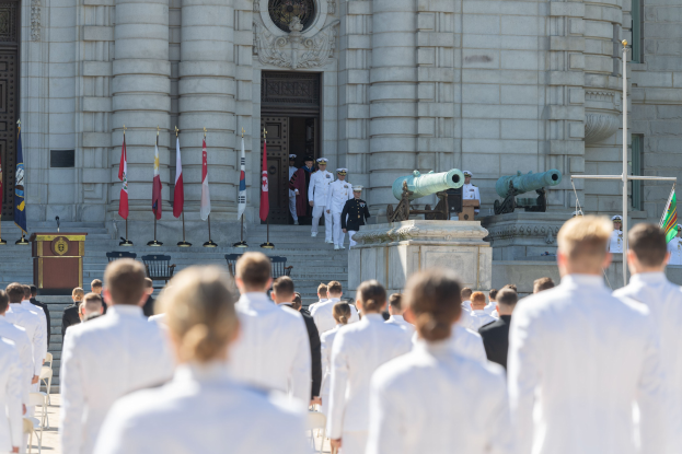 Gruppe von Menschen in weißen Marineuniformen auf einer Treppe vor einem Gebäude mit Säulen, Flaggen, einem Podium und Kanonen während einer Abschlussfeier.