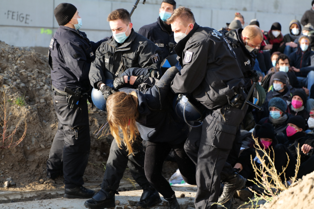Gruppe maskierter Personen in Kapuzenpullis, einige bewaffnet, die eine Frau umgeben, die von der Polizei eskortiert wird, mit städtischen Elementen wie Pflanzen, Steinen und einer Wand mit Protesttext im Hintergrund.