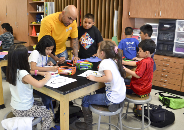 Eine Gruppe von Kindern sitzt um einen Klassentisch, während ein Mann vor ihnen steht, umgeben von Papieren, Stiften und Taschen, während Schränke und Regale mit Gegenständen im Hintergrund zu sehen sind.