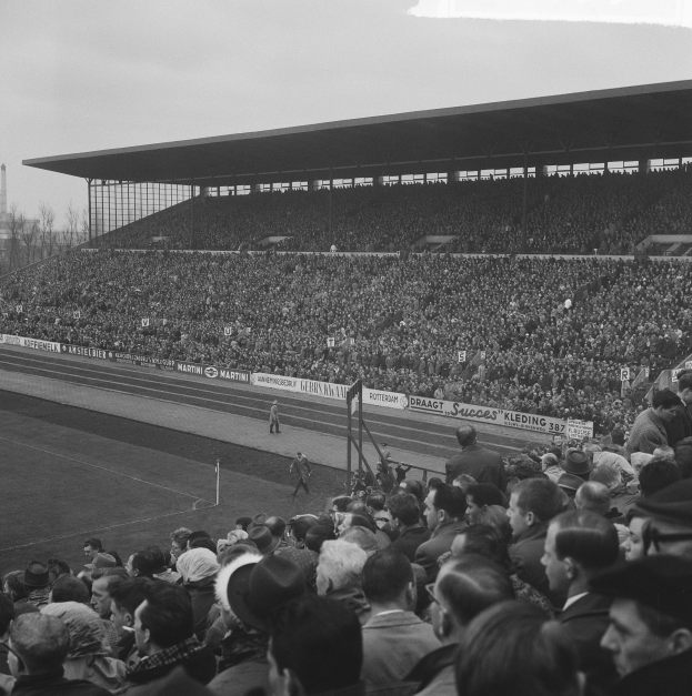 Schwarzes und weißes Foto einer vollen Stadion mit Zuschauern, die ein Fußballspiel verfolgen, mit Schildern, Pfählen, einem Schuppen, Bäumen, einem Turm und einem bewölkten Himmel.