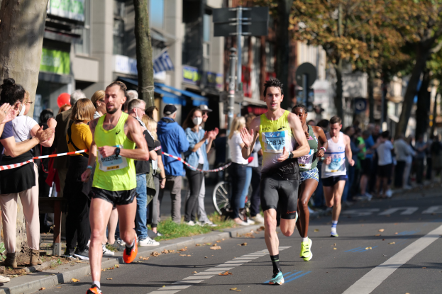 Gruppe von Läufern bei einem Marathon auf einer Stadtstraße, mit Zuschauern auf der linken Seite und Gebäuden, Bäumen und einem klaren blauen Himmel im Hintergrund.