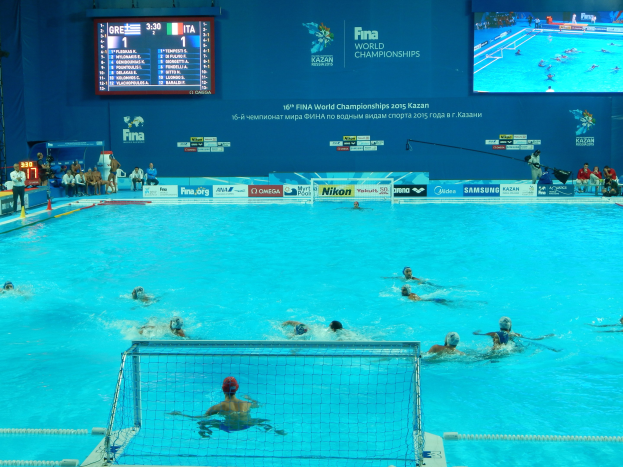 Gruppe von Athleten beim Wasserballspielen in einem Pool während der 2016 FINA-Weltmeisterschaften in Kazan, mit Zuschauern auf der linken Seite und einer Anzeigetafel mit Event-Branding im Hintergrund.