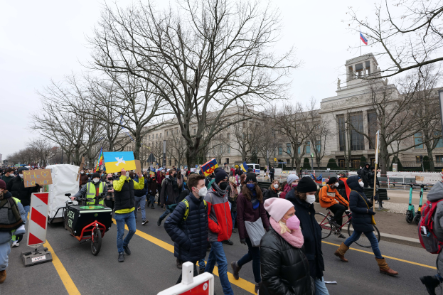Eine große Gruppe von Menschen nimmt an einer Protestdemo auf einer Straße in Washington, D.C. teil, einige halten Plakate und Schilder, andere fahren Fahrräder, und es gibt Schilder, Bäume und einen klaren blauen Himmel im Hintergrund.