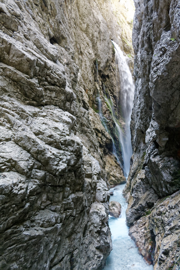 Ein kleiner Wasserfall ergießt sich über zerklüftete Felsen in einem steinigen Tal, umgeben von saftig grünen Hügeln unter strahlendem Sonnenschein.