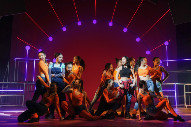 Eine Gruppe junger Tänzer in bunten Kostümen tanzt auf einer Bühne mit Geländern, die von bunten Lichtern beleuchtet wird, während das Publikum im Hintergrund jubelt.