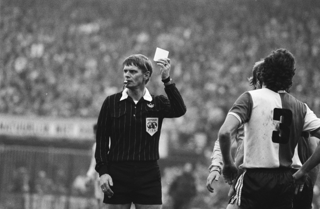 Ein Schiedsrichter in Schwarz-Weiß-Bild hält eine Rote Karte vor einer Menge von Menschen in Fußballuniformen, mit einem Zaun im Hintergrund.