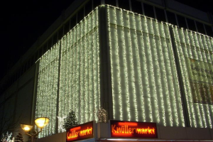 Ein Gebäude in Berlin, Deutschland, geschmückt mit Weihnachtslichtern, umgeben von Bäumen und Laternenmasten, mit Texttafeln in der Nähe und dem Himmel im Hintergrund.