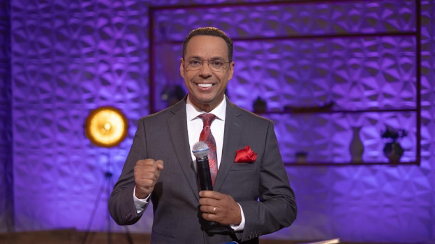 Stephen Colbert, Moderator von "The Daily Show with Trevor Noah", steht auf einer Bühne und hält ein Mikrofon in der Hand, trägt einen Anzug, eine Krawatte und eine Brille, während hinter ihm ein Scheinwerfer und ein Ständer zu sehen sind.