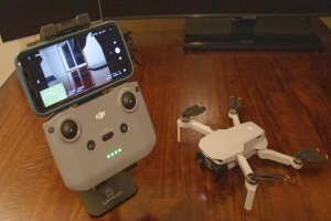 Ein DJI Mavic Air Fly More Combo Drohne, klein, faltbar, mit vier Rotoren und einer frontal montierten Kamera, steht auf einem Tisch mit einem verbundenen Mobiltelefon und einem Monitor im Hintergrund.