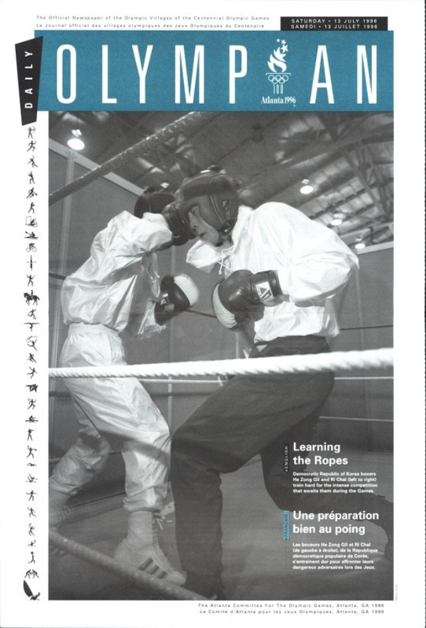Schwarz-weißes Foto von zwei Boxern in einem Boxring mit Helmen und Handschuhen, umgeben von Seilen unter beleuchteten Lichtern, mit 'Olympian Magazine'-Text oben und unten.