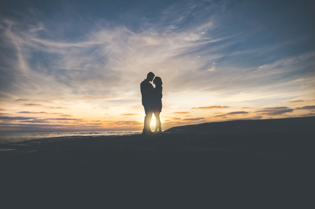 Silhouette eines Paares, das sich auf einem Strand bei Sonnenuntergang umarmt und küsst, mit leuchtendem orangefarbenem, pinkfarbenem und violetten Himmel und tiefblauem Wasser im Hintergrund.