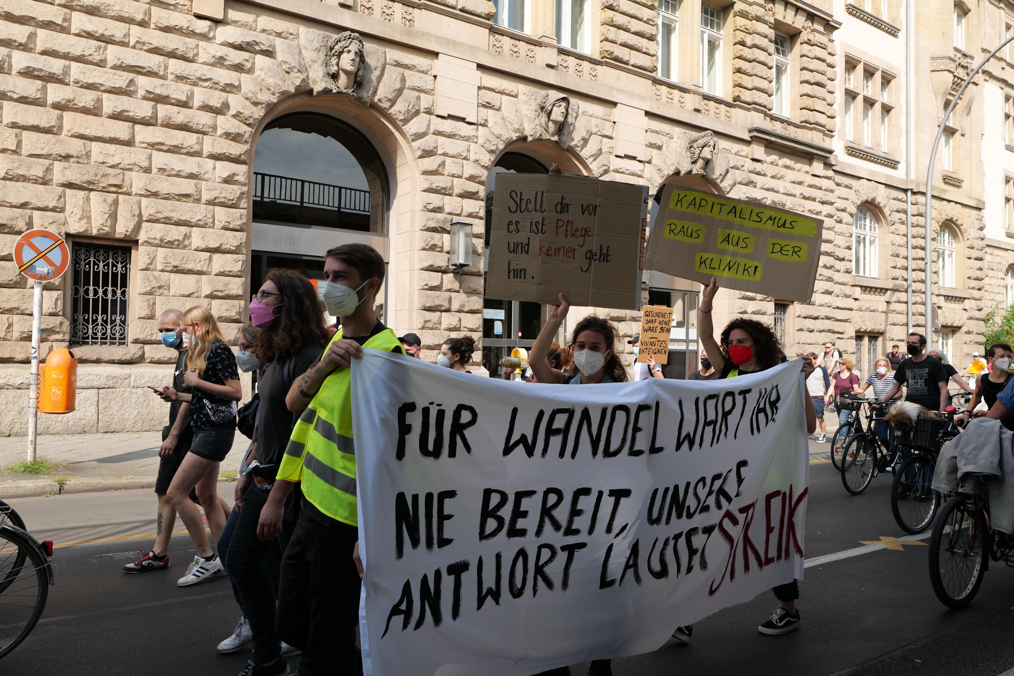 Eine Gruppe von Menschen marschiert bei einer Demonstration eine Straße entlang, einige halten Schilder und andere fahren Fahrräder, im Hintergrund ist ein Gebäude mit Fenstern, Bögen, Säulen und Skulpturen zu sehen.