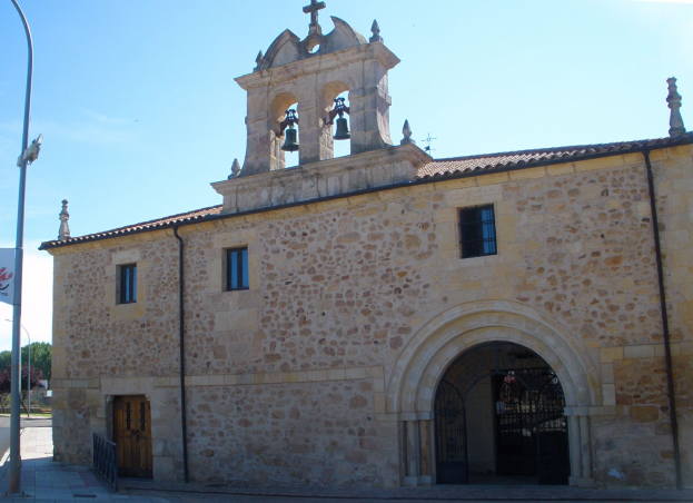 Ein altes Steingebäude mit einem zentralen Uhrenturm, umgeben von Straßenmasten, Lampen, Gittern, Bäumen und einem klaren blauen Himmel, mit Fenstern, Türen und Glocken am Turm.