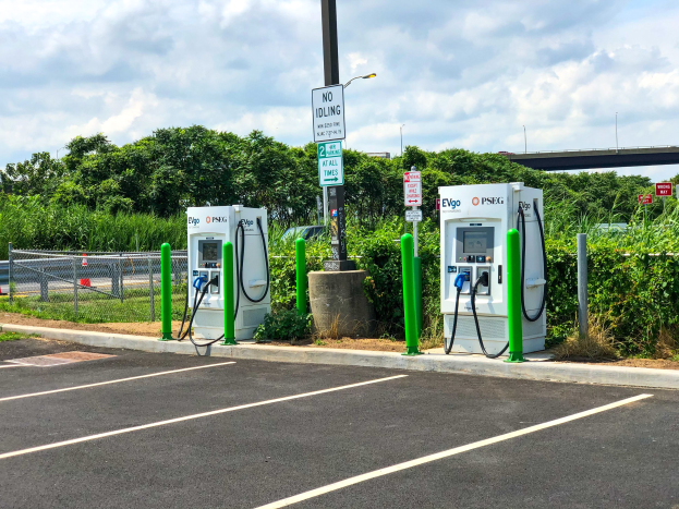 Elektroauto-Ladestation auf einem Parkplatz mit umgebender Infrastruktur, Vegetation und einer Brücke im Hintergrund bei bewölktem Himmel.