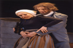 Ein Mann in einem Anzug mit ernstem Gesichtsausdruck und eine Frau mit einer Flasche stehen auf einer Bühne mit Text unten und einer Wand im Hintergrund.