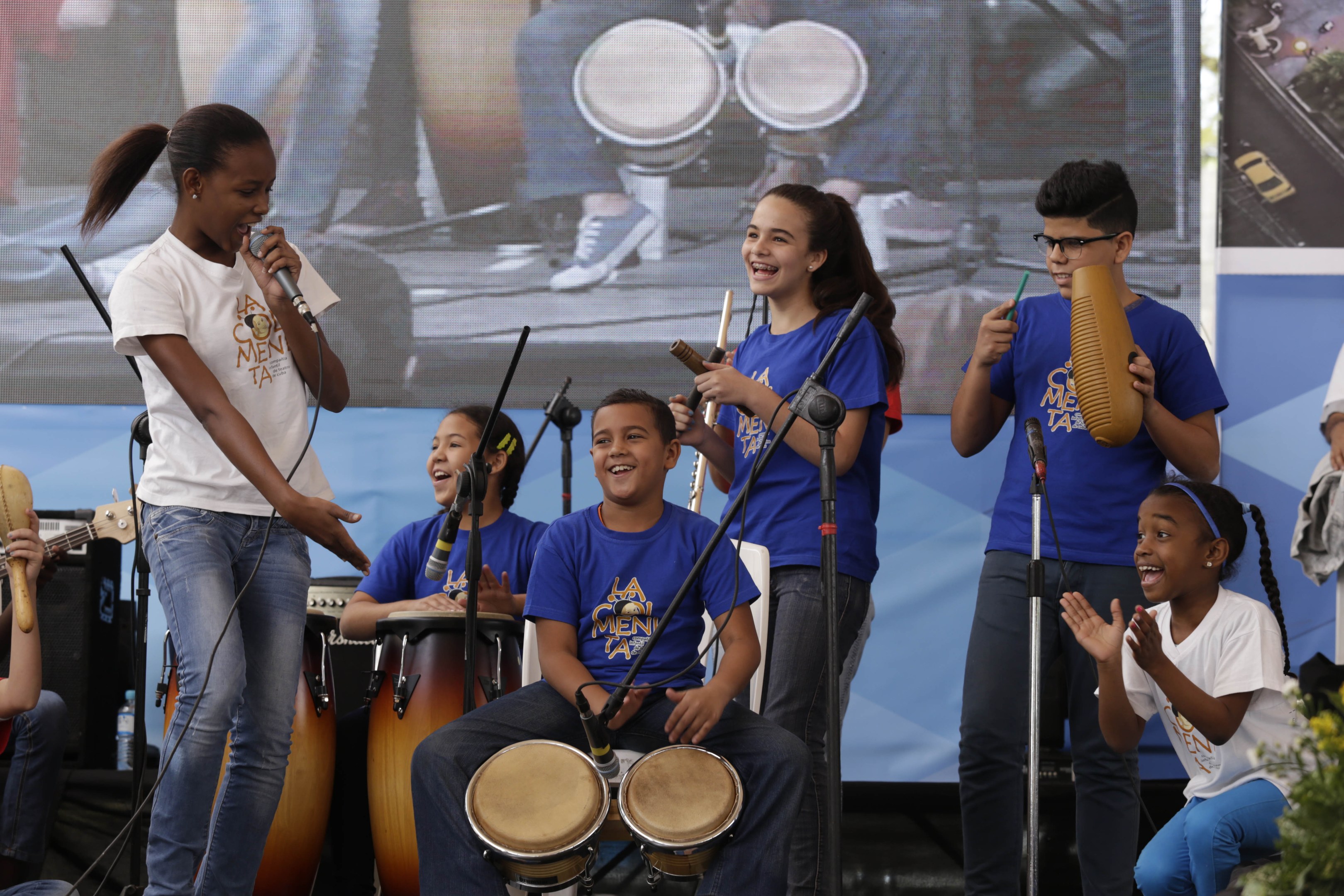 Gruppe von Kindern, die auf einer Bühne Musikinstrumente spielen, mit einer Frau in der Mitte, die ein Mikrofon hält, umgeben von Mikrofonständern und einem Banner im Hintergrund, mit Pflanzen auf der rechten Seite.