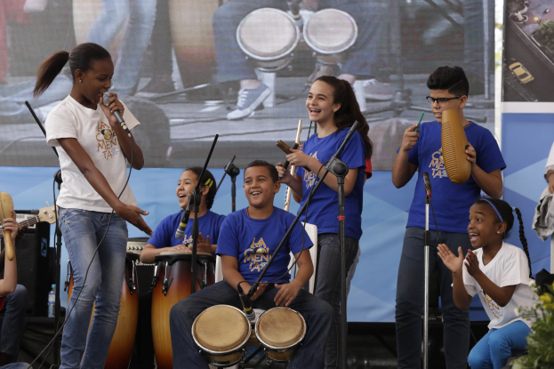 Gruppe von Kindern, die auf einer Bühne Musikinstrumente spielen, mit einer Frau in der Mitte, die ein Mikrofon hält, umgeben von Mikrofonständern und einem Banner im Hintergrund, mit Pflanzen auf der rechten Seite.