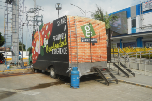 Lastwagen vor einem Gebäude mit einer Tafel, auf der "Teilen Sie eine positiv überladene Erfahrung" steht, zu sehen, mit Stufen, einem Zylinder auf dem Boden, Geländern, Strommasten mit Drähten, Laternenpfählen, Bäumen und einer wolkigen Himmel.