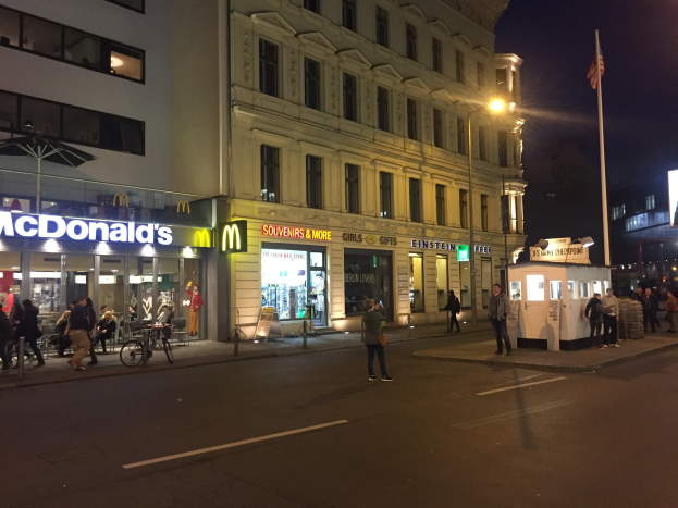 Gruppe von Menschen, die nachts eine Straße in Berlin entlanggehen, mit Gebäuden, Laternen, einer Fahne, Schaufenstern, Fahrrädern und einem sichtbaren Himmel.