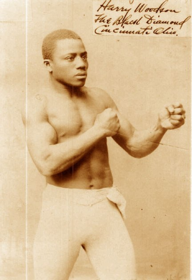 Ein Schwarz-Weiß-Foto eines Mannes in einem Boxring mit Handschuhen und Shorts, der mit konzentrierter Miene und erhobenen Armen bereitsteht.