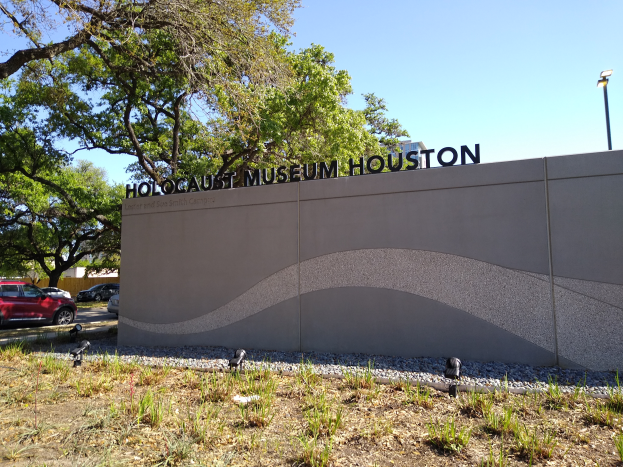 Das Holocaust-Museum in Houston, Texas, mit einer textbedeckten Wand, Steinen, Gras, Pflanzen, einer Baumgruppe, Fahrzeugen auf der Straße, einem Straßenschild und einem bewölkten Himmel.