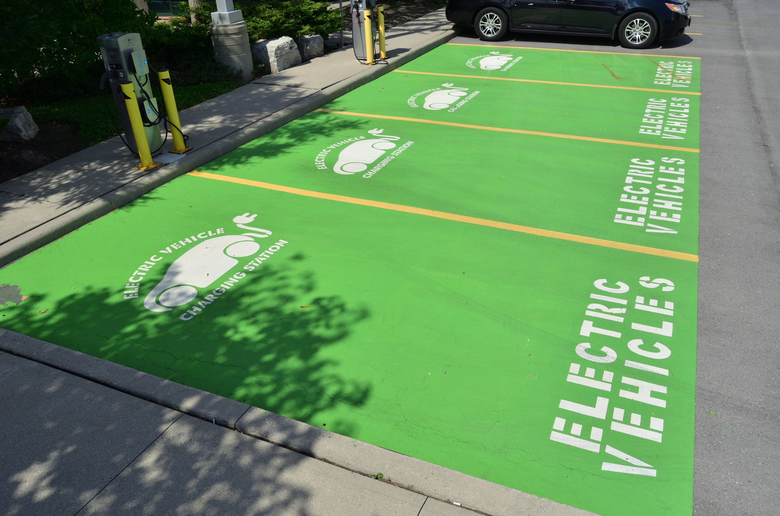 Elektroauto auf einem Ladeplatz mit grüner Bodenmatte mit Text und Bildern in einer Parkanlage mit Bäumen und Pfählen.