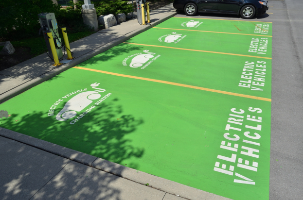 Elektroauto auf einem Ladeplatz mit grüner Bodenmatte mit Text und Bildern in einer Parkanlage mit Bäumen und Pfählen.