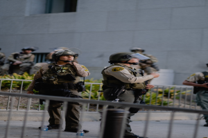 Eine Gruppe uniformierter Polizisten, einige bewaffnet, steht in einer Reihe vor einem Gebäude, mit einem Metallzaun im Vordergrund und Pflanzen und einer Wand im Hintergrund.