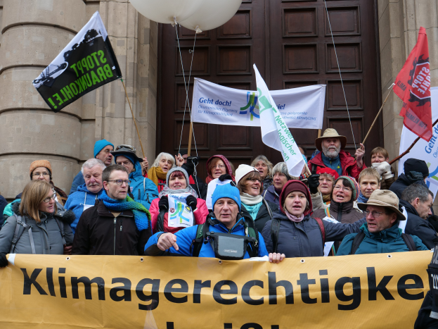Eine Gruppe von Menschen vor einem Gebäude mit Säulen und einer Tür, die ein Transparent mit Text, Fahnen und Luftballons halten, nimmt an einem Klimastreik in Deutschland teil.