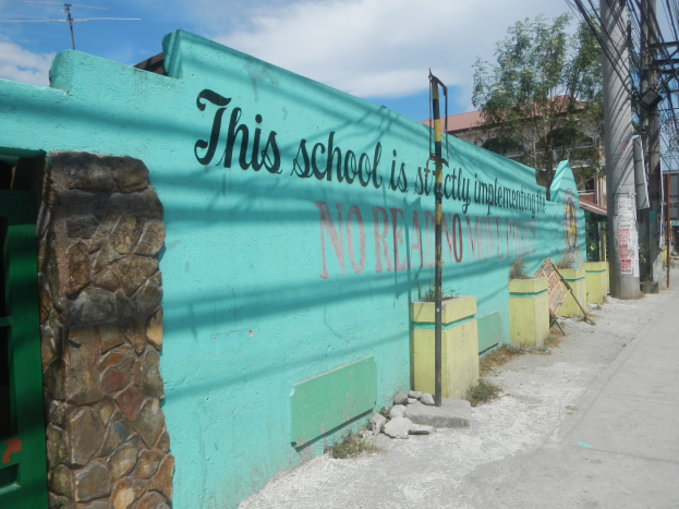 Grüne Wand mit Graffiti, das 'Diese Schule ist keine Stadt' liest, umgeben von Gebäuden, Bäumen, Strommasten, Drähten und anderen städtischen Elementen unter einem bewölkten Himmel.