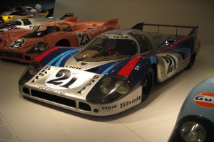 Ein Porsche 917K Le Mans Rennauto wird in einem Museum ausgestellt, umgeben von anderen Sportwagen und Menschen, mit einer Wand im Hintergrund und einem Blatt Papier mit Text links.