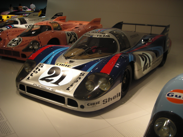 Ein Porsche 917K Le Mans Rennauto wird in einem Museum ausgestellt, umgeben von anderen Sportwagen und Menschen, mit einer Wand im Hintergrund und einem Blatt Papier mit Text links.