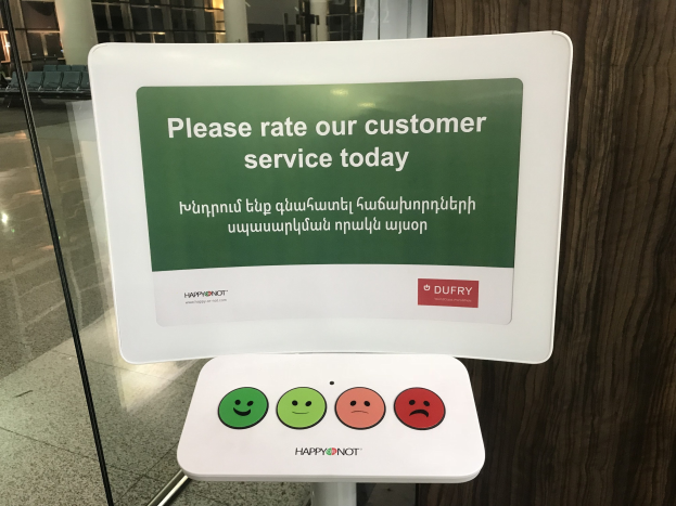 Ein Schild mit der Aufschrift "Bewerten Sie unseren Kundenservice heute" steht vor einer Glastür mit einer hölzernen Wand rechts daneben und einem Boden darunter, mit Stühlen, Säulen und Lampen im Hintergrund.