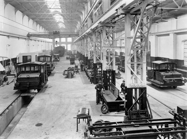Ein Schwarz-Weiß-Foto eines Fabrikinneren mit mehreren Zügen auf dem Boden, Arbeitern, Metallstangen, Deckenlampen und Wandfenstern.