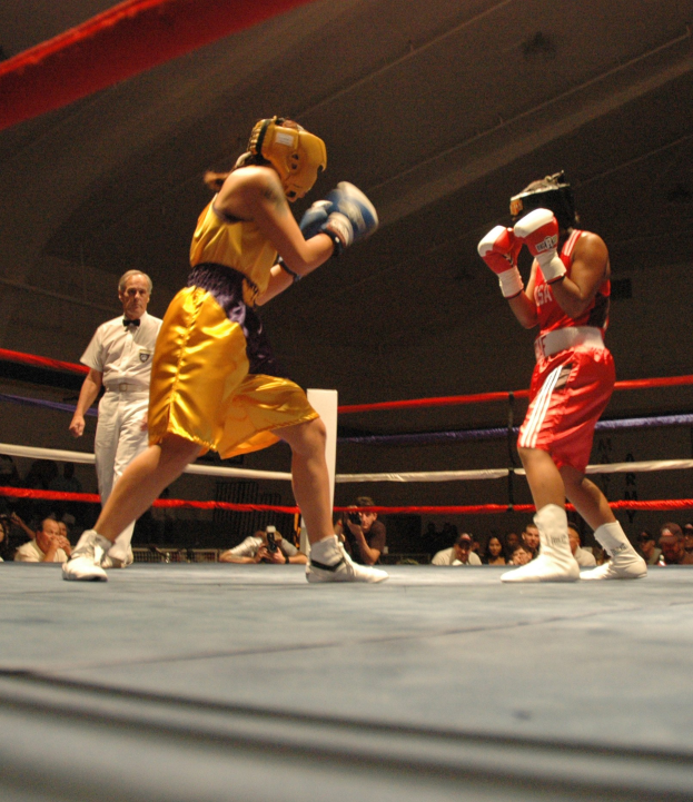 Zwei Boxer kämpfen im Ring mit einem Schiedsrichter und Zuschauern, einige mit Kameras, unter einer sichtbaren Decke.