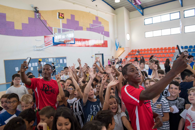 Eine Gruppe von Kindern steht vor einem Basketballfeld, einige halten Mobiltelefone, mit einer Tafel, Uhr, Torstange, Basketballkorb, Deckenleuchten, Stühlen und Fenstern im Hintergrund.