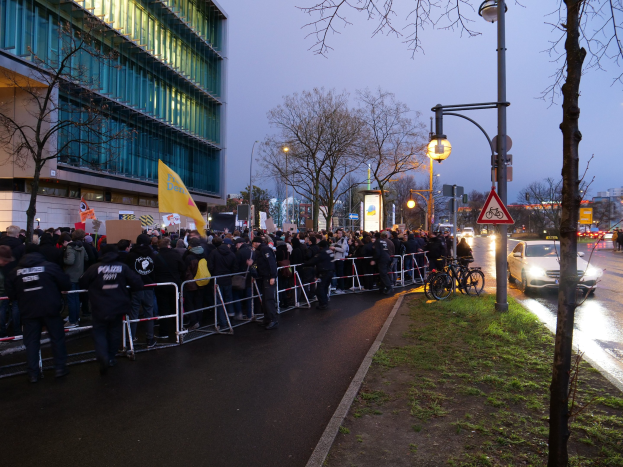 Eine große Gruppe von Menschen mit Protestschildern steht vor einem Gebäude in Berlin, mit Barrikaden, Fahrrädern, Laternenmasten, Schildern, Bäumen und Gras auf dem Boden.