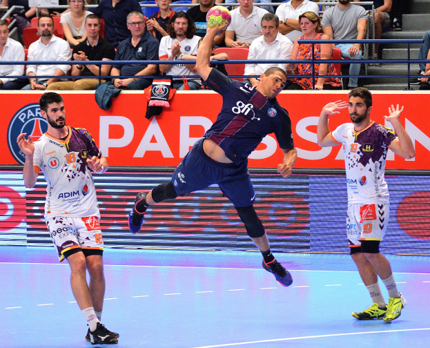 Mannergruppe spielt Handball auf einem Platz mit einem Ball im Spiel, umgeben von Zuschauern und verstreuten Kleidungsstücken, mit einem Banner für das Futsal-Weltmeisterschaftsspiel 2015 zwischen Paris Saint-Germain und Olympique Lyon im Hintergrund.
