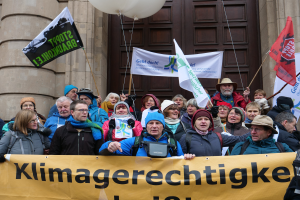 Eine Gruppe von Menschen vor einem Gebäude mit Säulen und einer Tür, die ein Transparent mit Text, Fahnen und Luftballons halten, nimmt an einem Klimastreik in Deutschland teil.