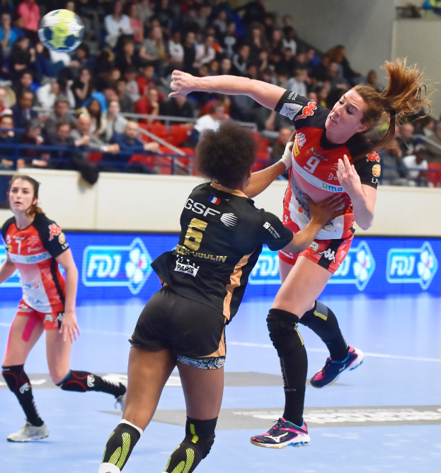 Eine Gruppe von Frauen spielt Handball auf einem Platz mit einem Ball in der Luft, während Zuschauer von Stühlen, Geländern und einer Wand im Hintergrund zusehen.