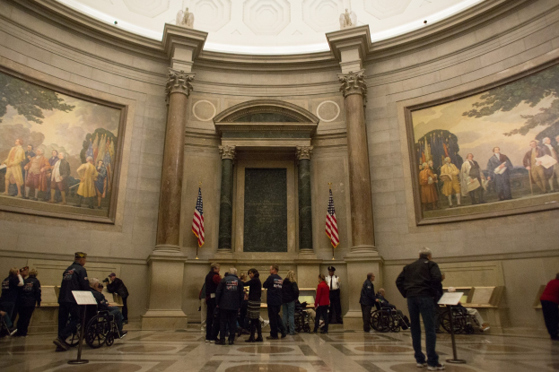 Eine Gruppe von Menschen vor einem Gebäude mit Kuppel, einige im Rollstuhl, andere stehend, mit Ständen mit Papieren, Wänden mit Fotorahmen und Fahnenmasten, und einer Decke mit Statuen.