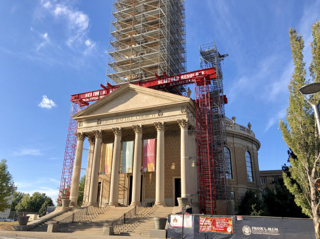 Großes Gebäude im Bau mit Gerüst, mit Säulen, Treppen mit Geländern, Bannern, einem Laternenmast, Bäumen, Fahrzeugen auf der Straße und einem bewölkten Himmel.