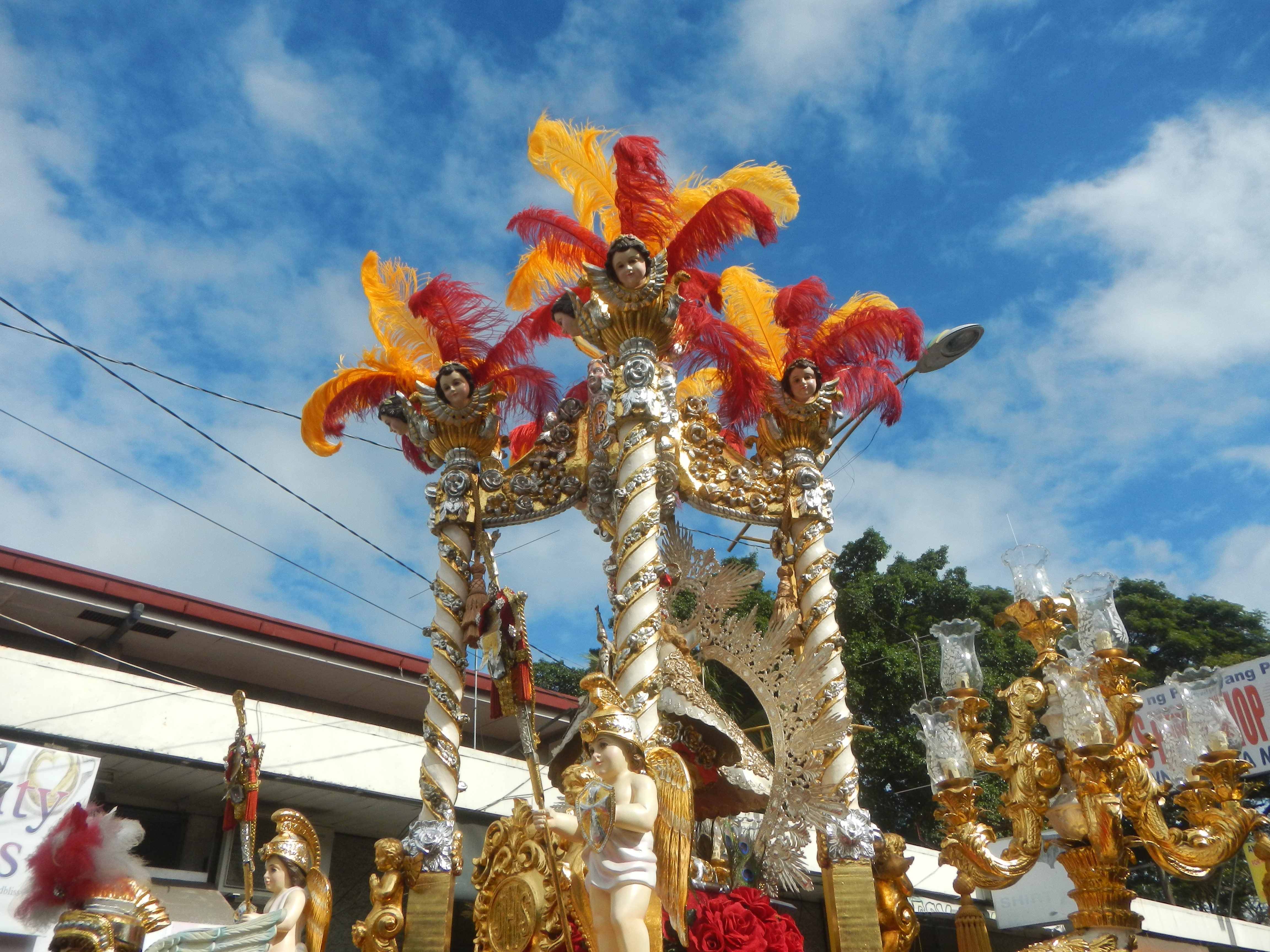 Ein Karnevalswagen, geschmückt mit goldenen und roten Federn, der Menschen trägt, wird in einem Umzug mit einem Gebäude, Bäumen, Drähten und einem Banner in der Nähe, unter einem sichtbaren Himmel gezeigt.
