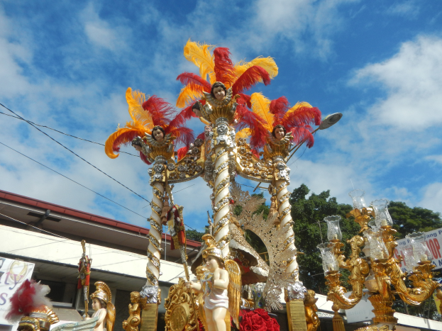 Ein Karnevalswagen, geschmückt mit goldenen und roten Federn, der Menschen trägt, wird in einem Umzug mit einem Gebäude, Bäumen, Drähten und einem Banner in der Nähe, unter einem sichtbaren Himmel gezeigt.