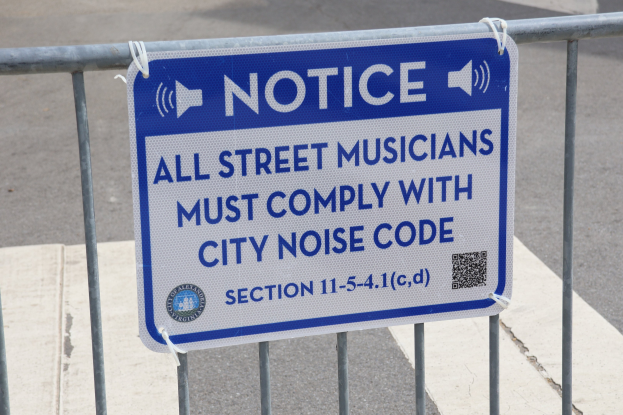 Anschlagtafel an einem Zaun mit einem Schild über die Einhaltung des Lärmcodes für Straßenmusiker, mit einer Straße im Hintergrund.