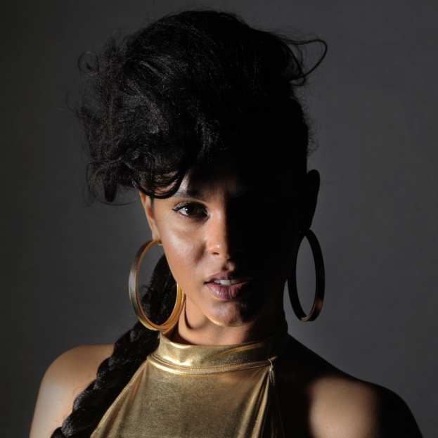 Eine Frau in einem goldenen Kleid und großen Ohrringen steht selbstbewusst vor einer Wand, ihr glatter Bob-Haarschnitt und das natürliche Make-up unterstreichen ihren entschlossenen Gesichtsausdruck, während sie direkt in die Kamera blickt.