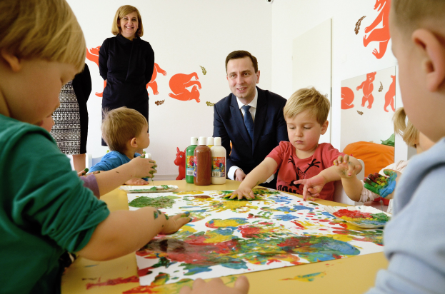 Kinder beim Malen an einem Tisch mit Farbe an den Händen, Papieren und Flaschen, während eine Frau im Hintergrund mit Gemälden an der Wand lächelt.